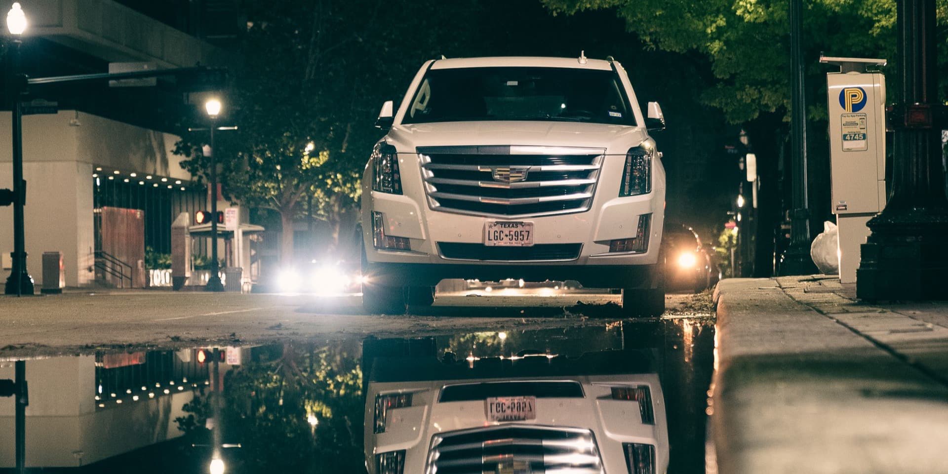 White Cadillac Escalade parked on a downtown street at night, mirrored in the wet pavement